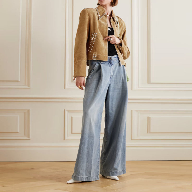 A model wearing a tan suede jacket with silver studs, a black top, and light blue wide-leg denim jeans, standing in a room with cream paneled walls and a light wood floor.