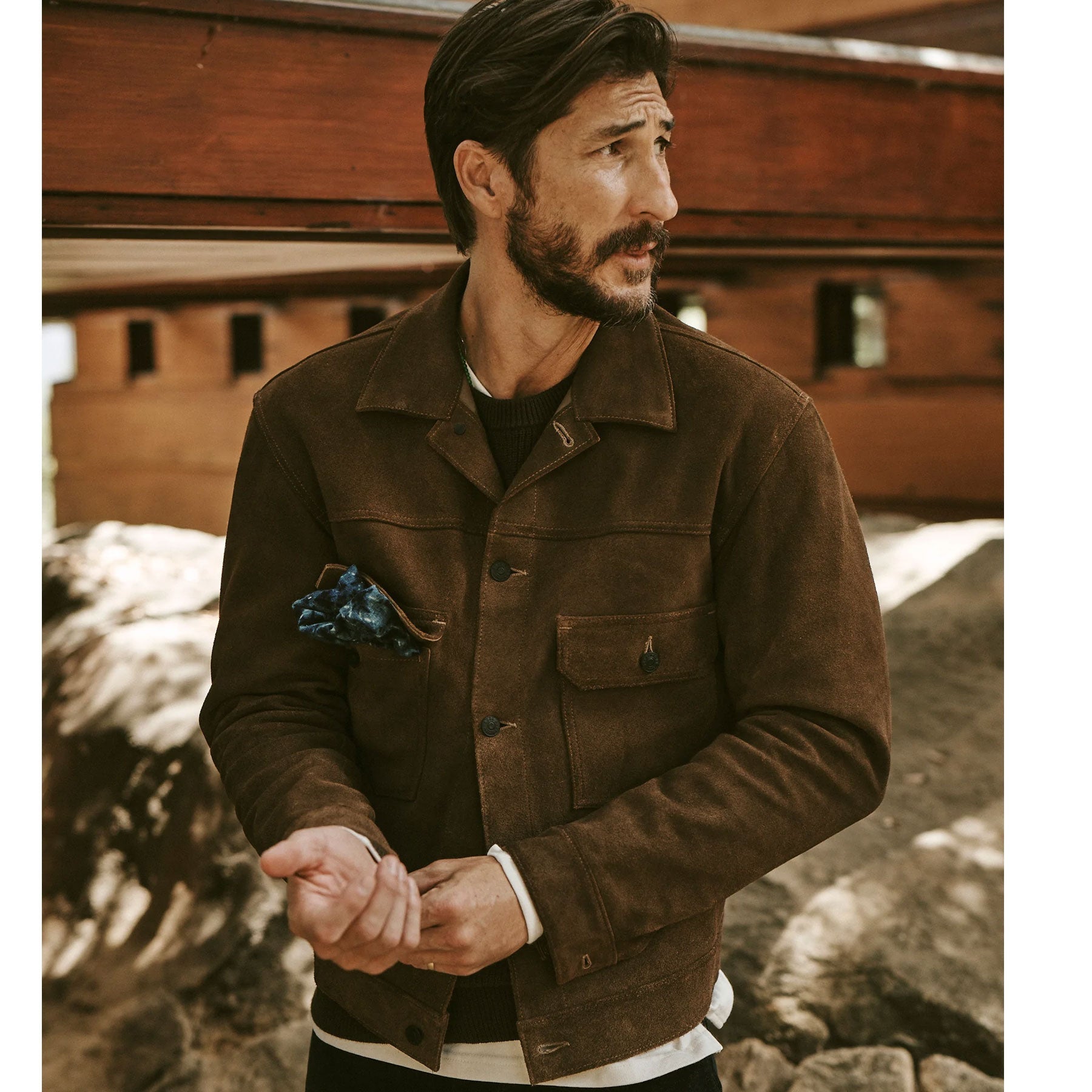 Man wearing a brown jacket with a wooden building in the background