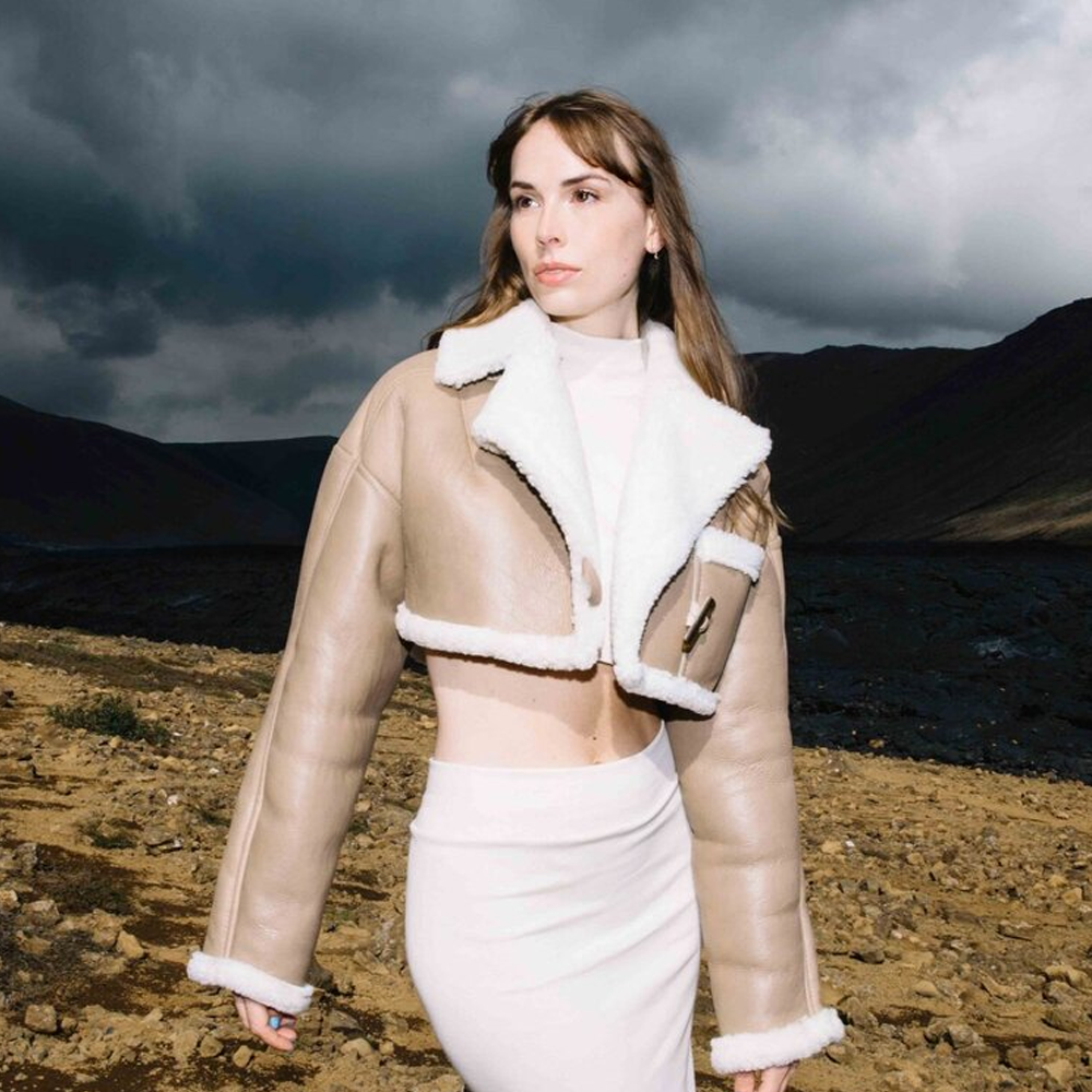 Woman wearing a beige puffer jacket with white fur trim in a barren landscape under a stormy sky.