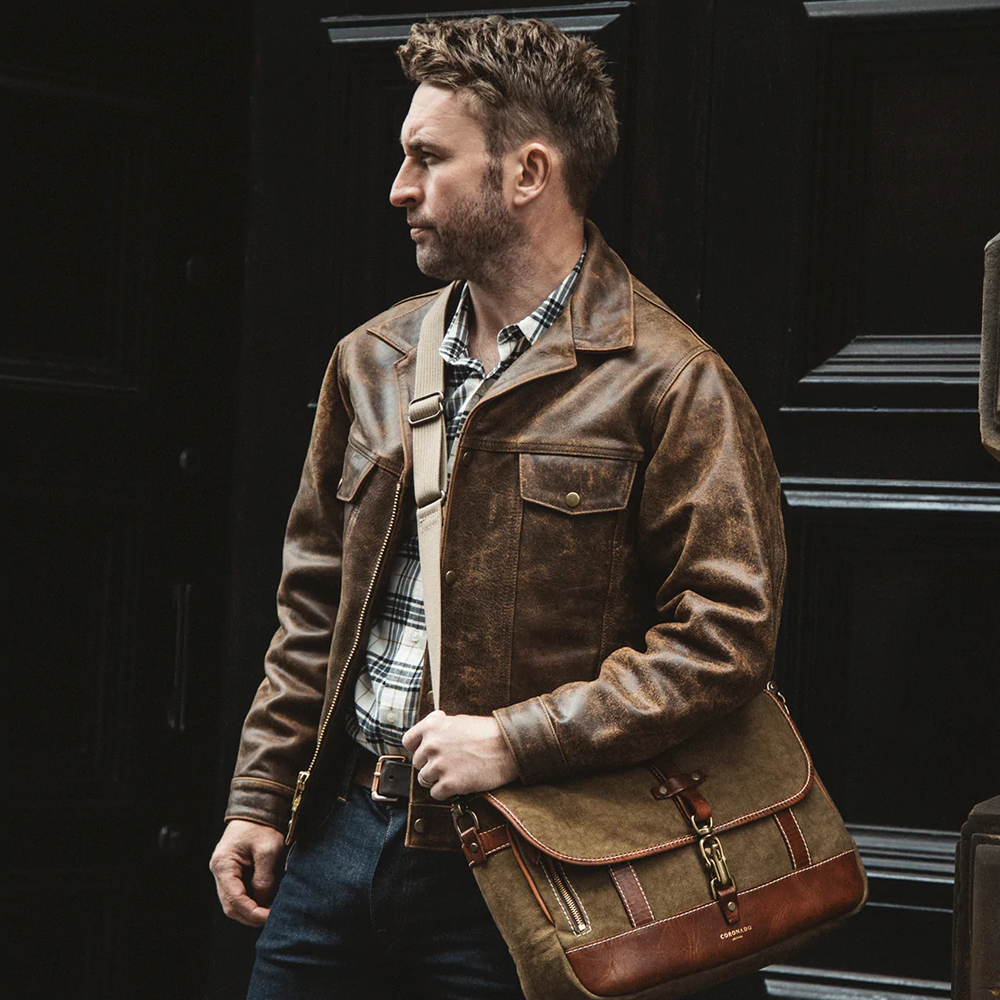 Man wearing a brown leather jacket holding a matching bag against a dark background