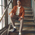 Man in an orange jacket and brown pants sitting on a staircase.