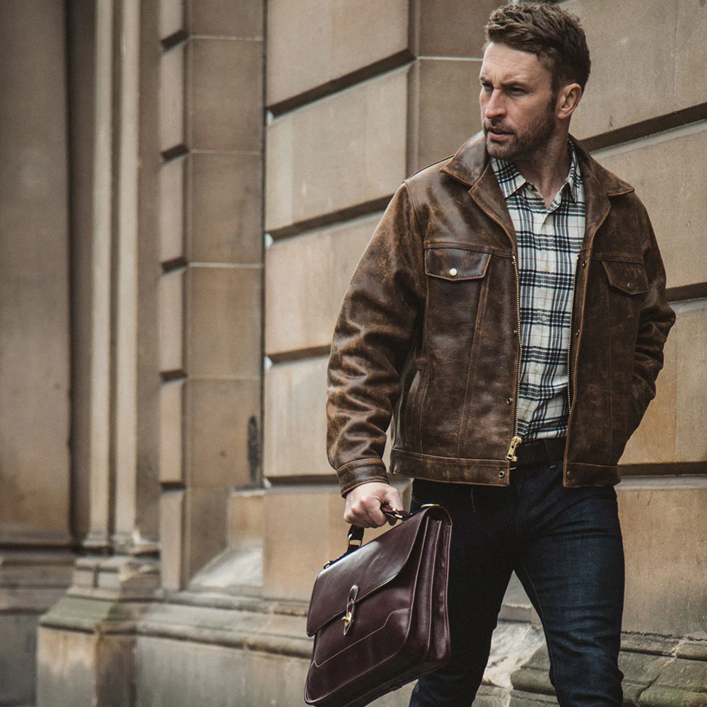 Man in a brown leather jacket holding a brown leather briefcase against a stone building background