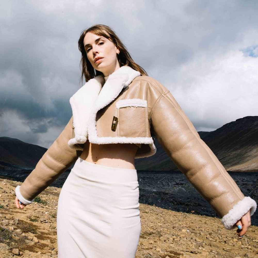 Woman wearing a beige shearling jacket with white fur trim against a mountainous landscape.