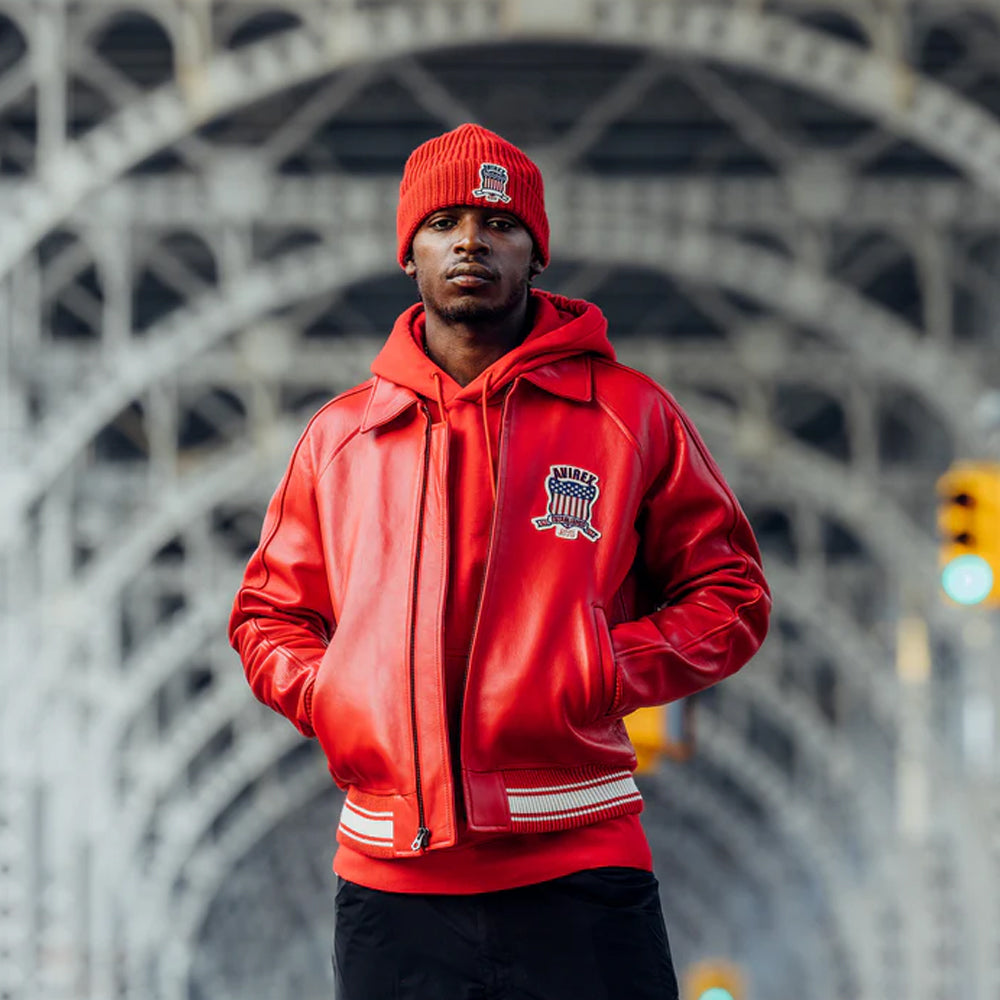 A man wearing a bright red leather jacket, matching red beanie, and black pants stands in front of a large steel arch bridge. The jacket has a zip-up front, ribbed trim with white stripes, and a crest patch on the left chest.