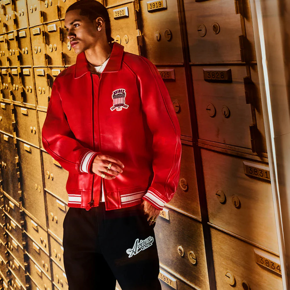 A man wearing a red leather varsity-style jacket with white striped ribbed cuffs and hem, and a white graphic on the left chest, standing in front of a row of metallic bank vaults.