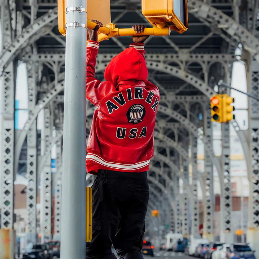 A person seen from behind wearing a bright red hooded jacket with 'AVIREX USA' printed on the back, standing on a city bridge and touching a yellow traffic light. The jacket has white stripes and a crest patch on the back.