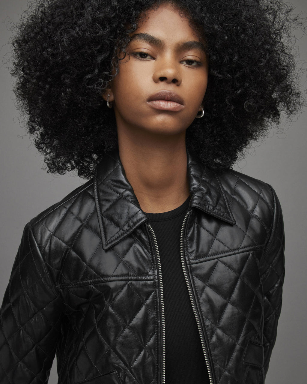 A close-up portrait of a model with voluminous black curly hair wearing a black quilted leather jacket over a black top, set against a solid gray studio background.