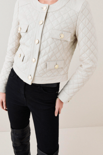 A close-up of a person wearing a light beige quilted jacket with gold-tone buttons and black pants, cropped from the neck down against a plain light background.