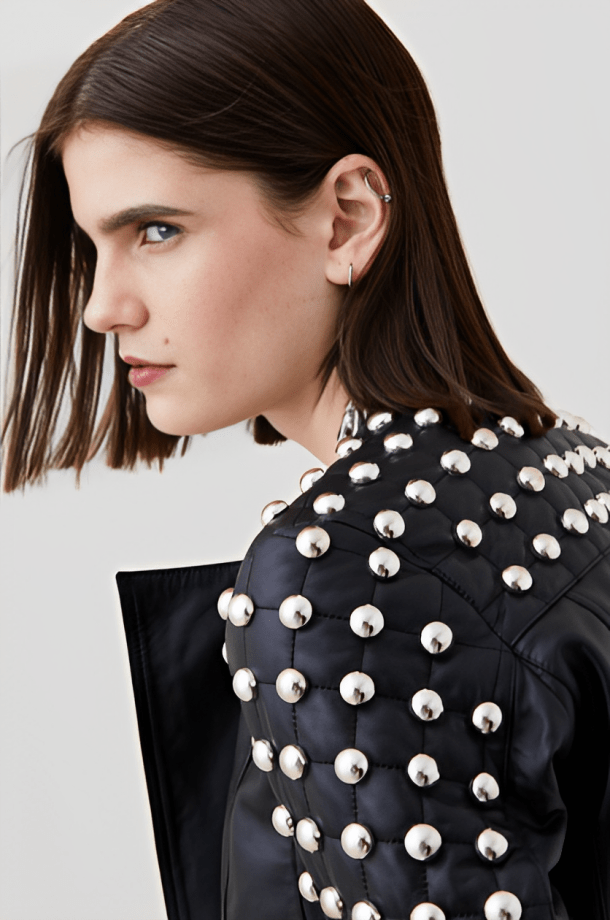 Close-up side profile of a model wearing a black quilted jacket heavily embellished with large silver dome studs, against a light gray studio background.