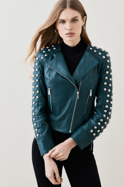 A woman wearing a dark teal leather moto jacket with silver studs on the shoulders and sleeves, styled over a black turtleneck and black pants, against a light gray studio background.