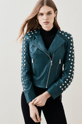 A woman wearing a dark teal leather moto jacket with silver studs on the shoulders and sleeves, styled over a black turtleneck and black pants, against a light gray studio background.