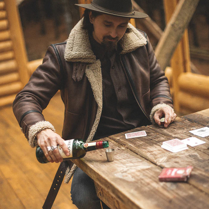 Man in a brown leather jacket and hat pouring a drink at a wooden table with playing cards.