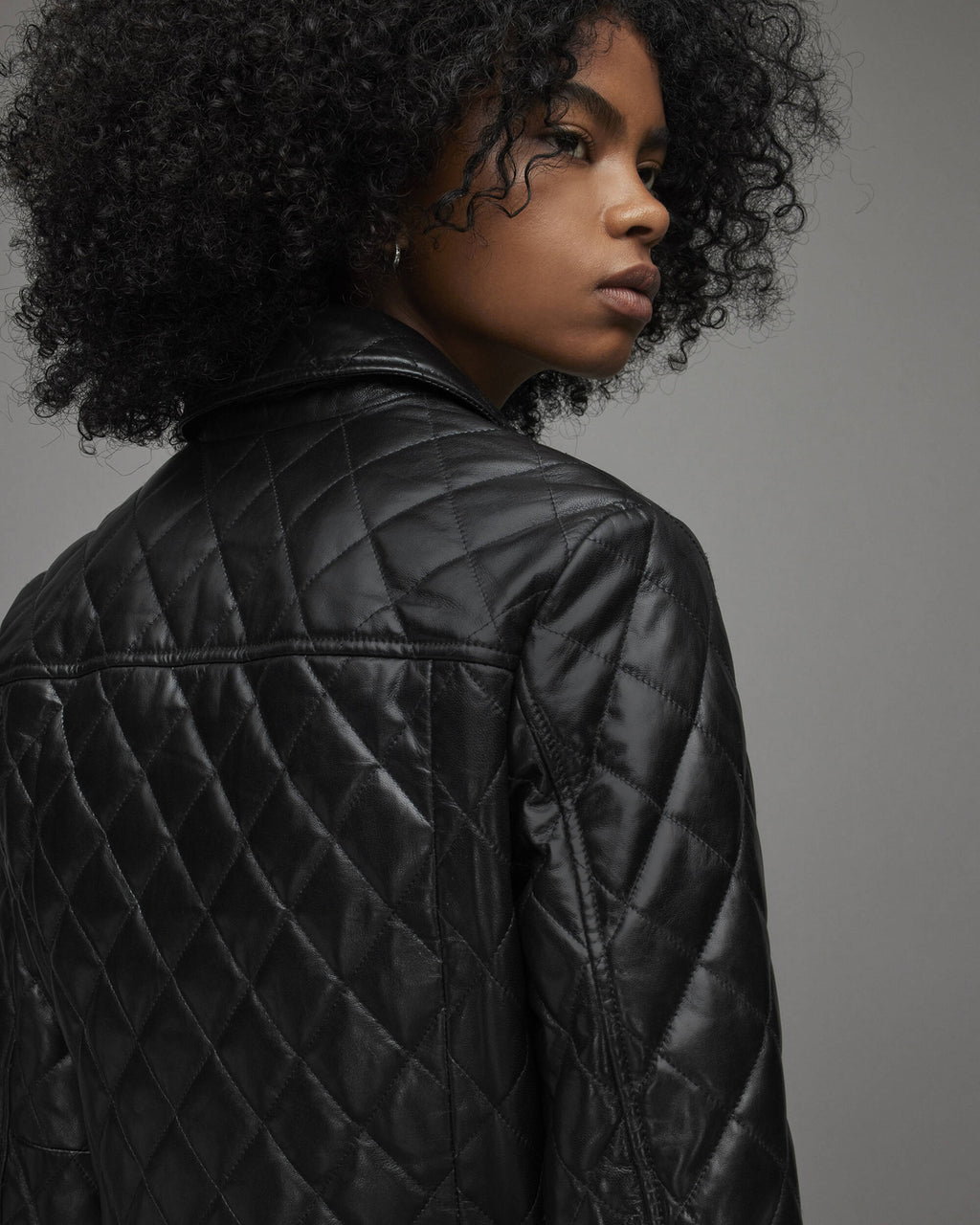 A close-up side profile of a model with curly black hair wearing a black quilted leather jacket against a solid gray studio background.