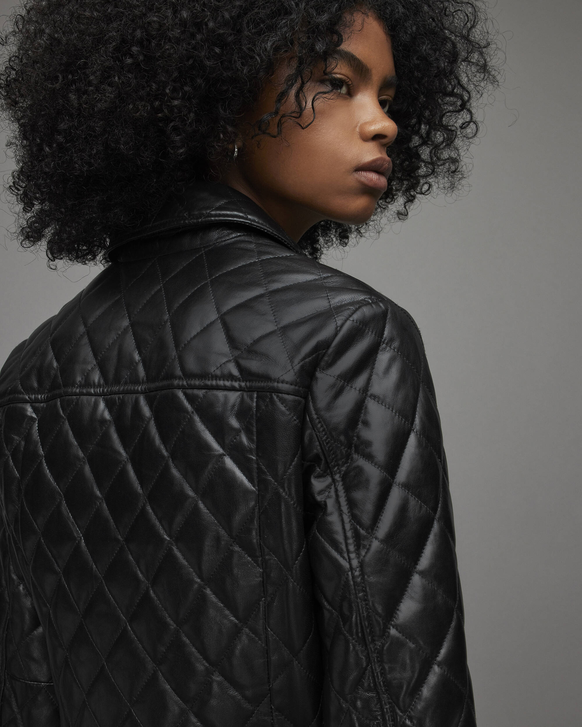 A close-up side profile of a model with curly black hair wearing a black quilted leather jacket against a solid gray studio background.
