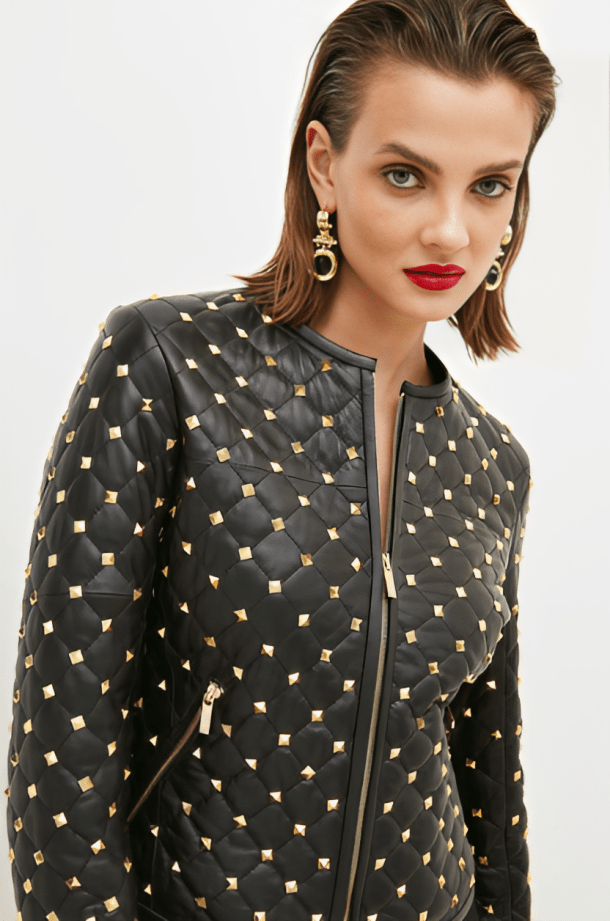 A close-up of a model wearing a black quilted jacket embellished with gold pyramid studs, set against a plain light gray background.