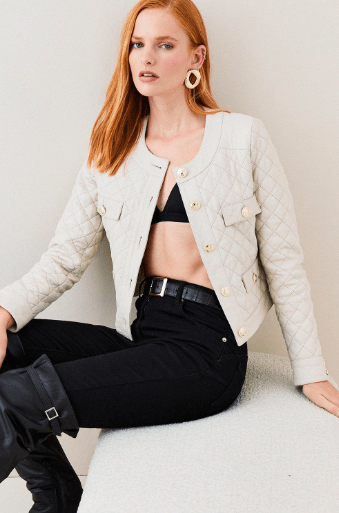 A red-haired model wearing a light beige quilted jacket with gold buttons, black pants, and black boots, sitting against a light gray background.