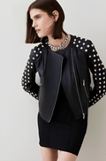 A model wearing a black moto jacket with silver studs on the sleeves and collar, styled over a black dress and silver chain necklace, against a light gray studio background.