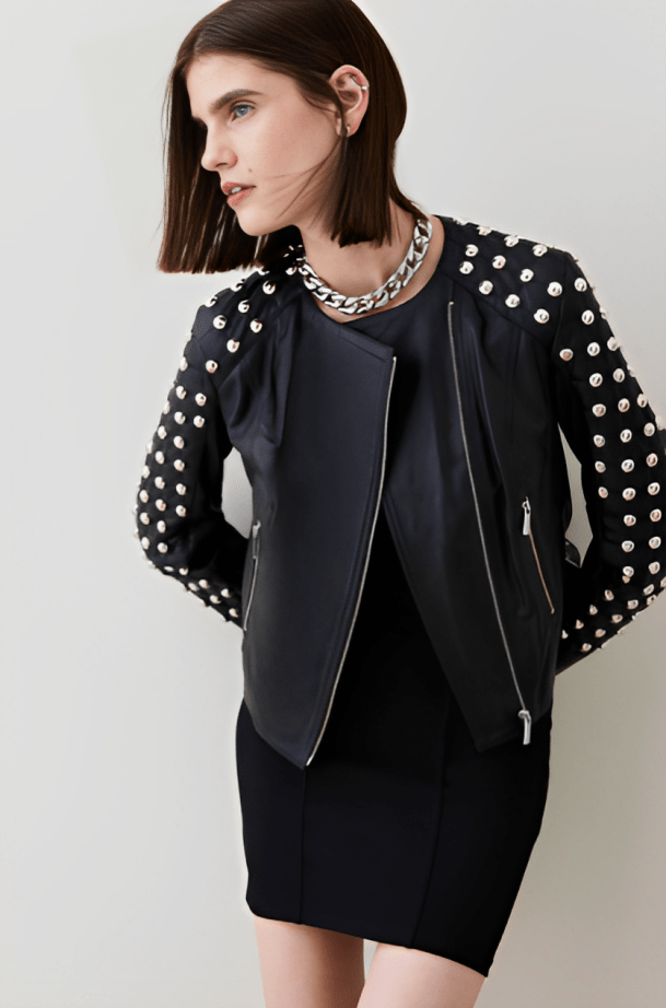 A model wearing a black moto jacket with silver studs on the sleeves and collar, styled over a black dress and silver chain necklace, against a light gray studio background.