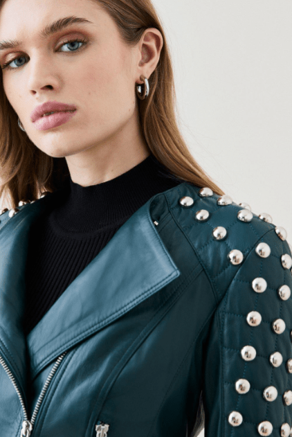 Close-up of a woman wearing a dark teal leather moto jacket with silver studs on the shoulders and sleeves, layered over a black ribbed turtleneck.
