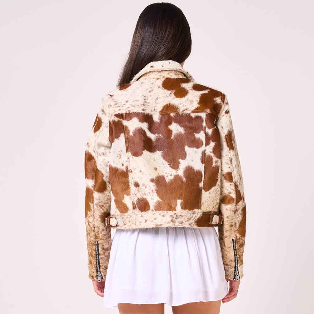 A woman viewed from the back wearing a cropped cow-print jacket with brown and cream patches, paired with a white pleated skirt, against a plain light background.