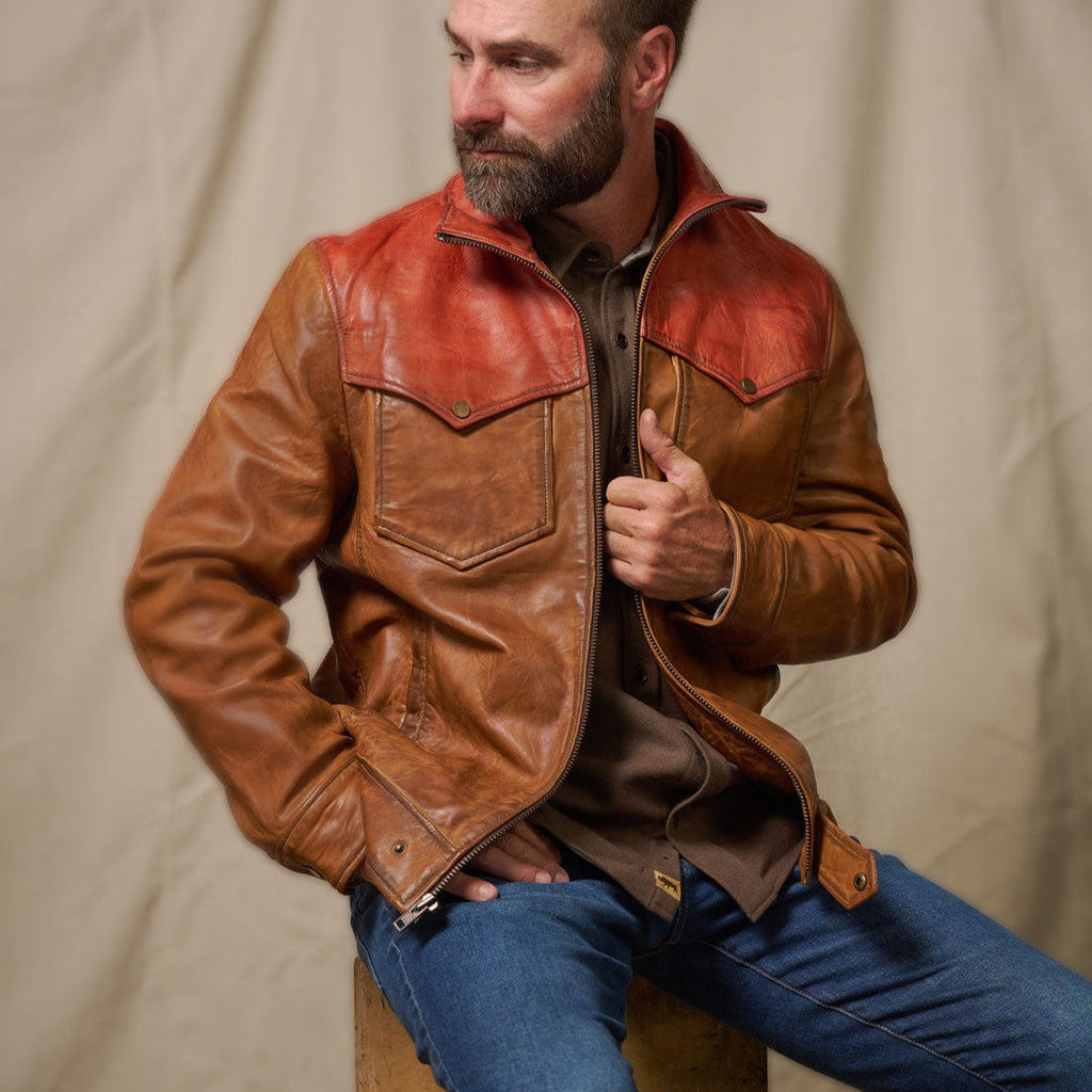 Man wearing a brown leather jacket with red accents, sitting on a stool against a plain background.