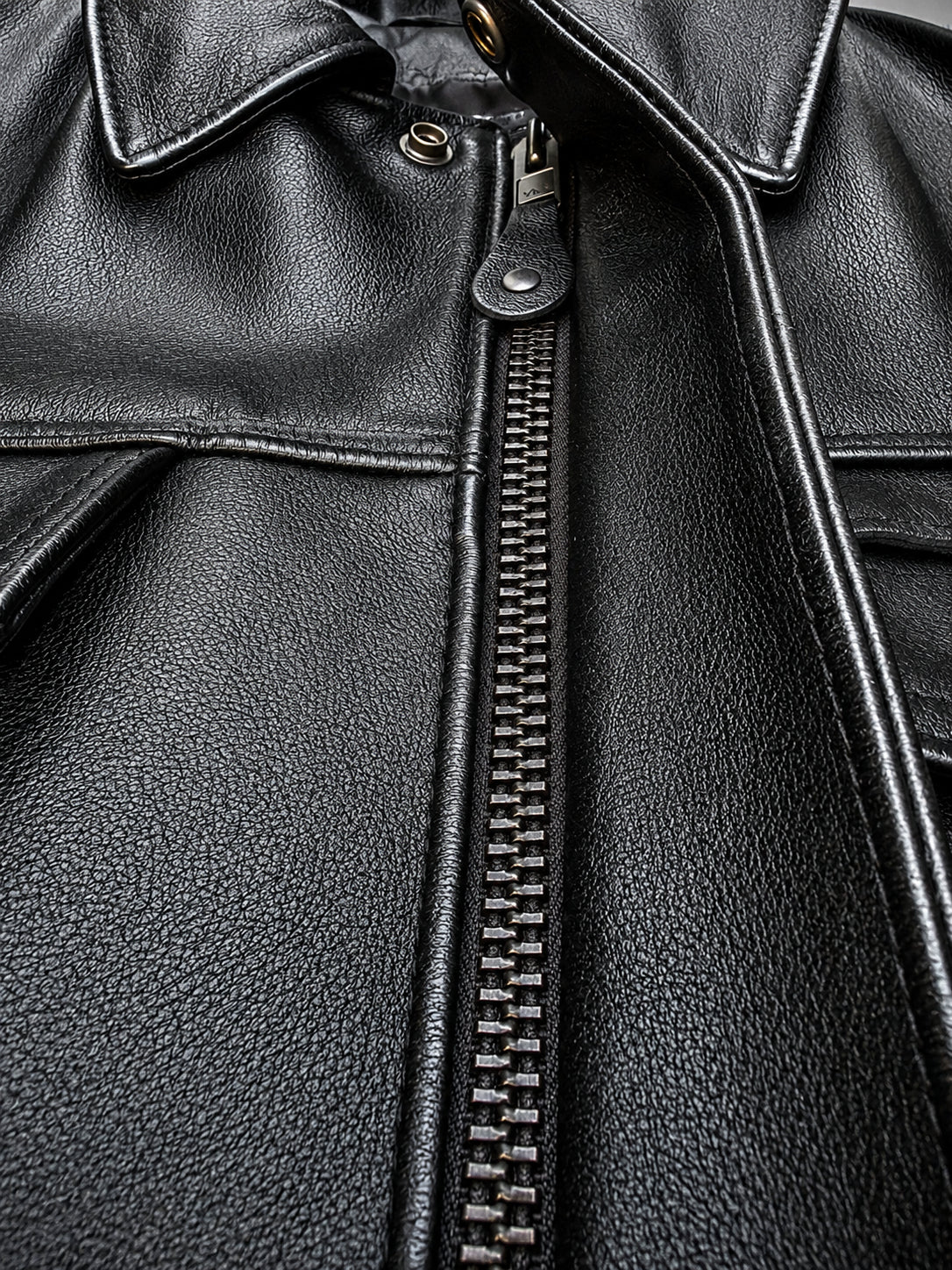 Close-up of a black pebbled leather jacket showing the front zipper, collar, and stitched details.