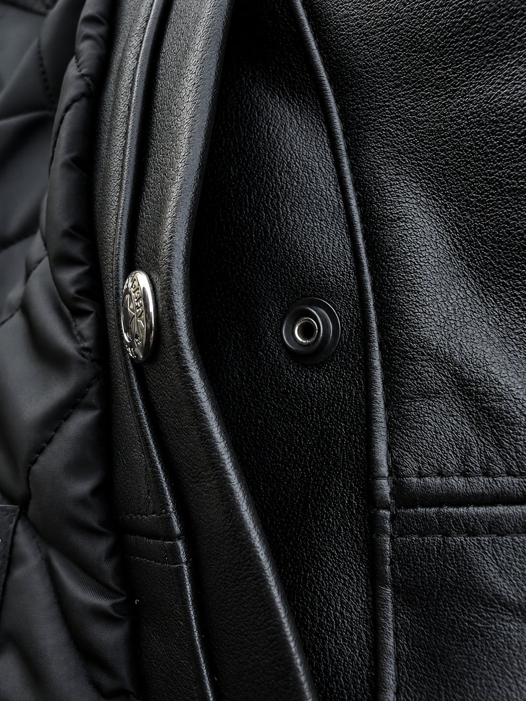 Close-up detail of a black jacket showing textured leather-like fabric, quilted lining, and silver-tone snap hardware.