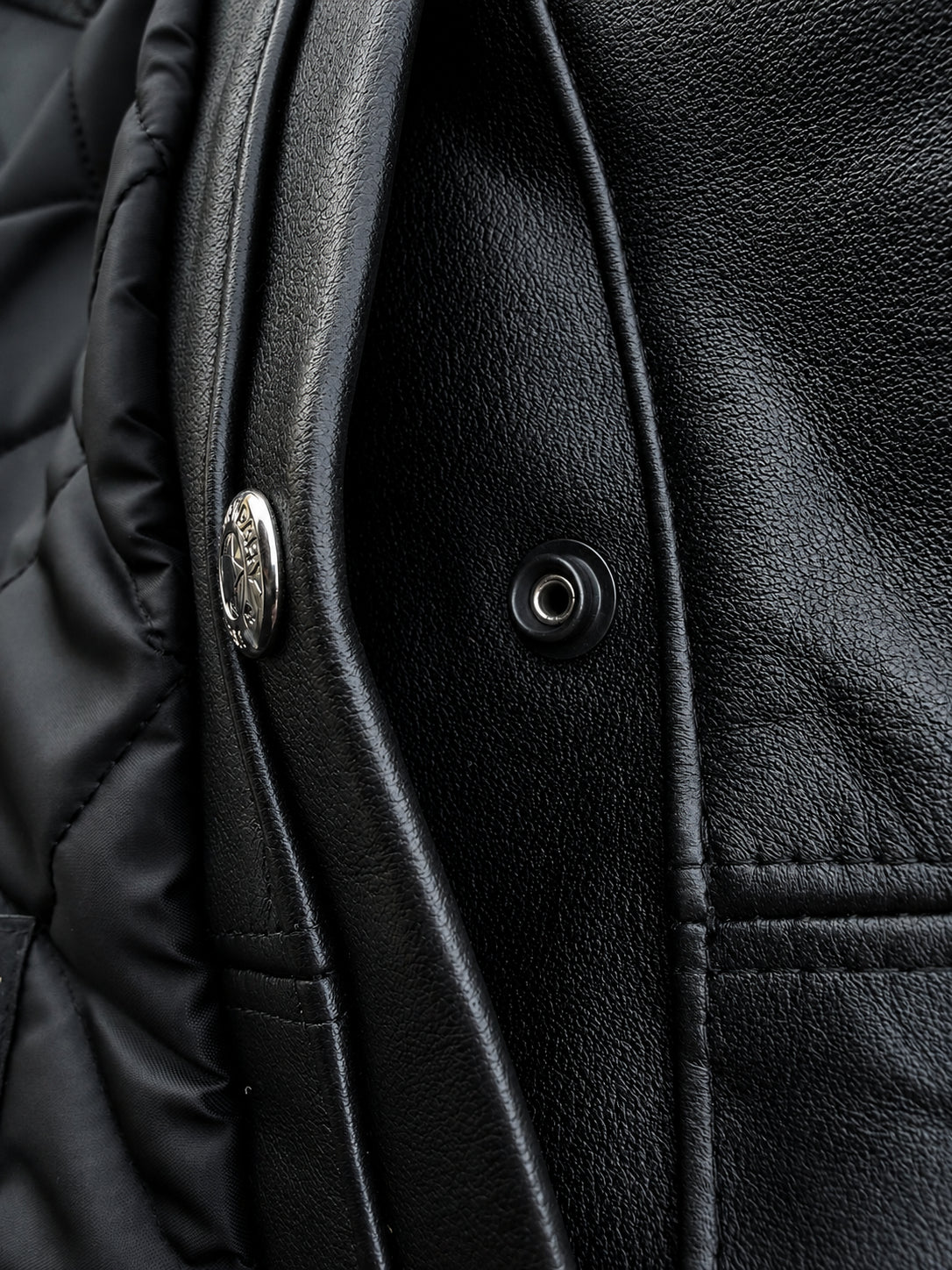 Close-up detail of a black jacket showing textured leather-like fabric, quilted lining, and silver-tone snap hardware.
