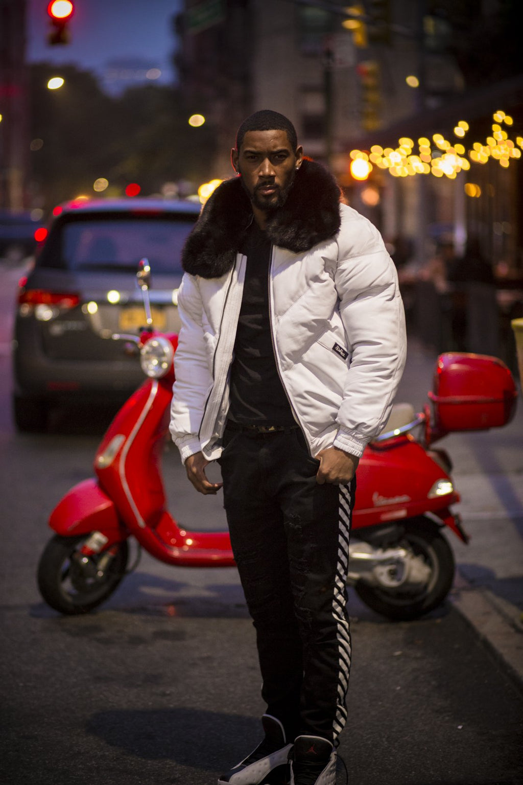 Man in a white jacket with fur trim standing next to a red scooter on a city street at night.