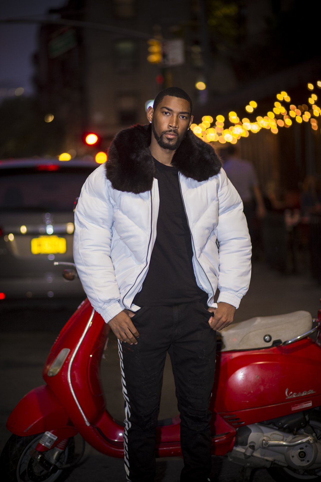 Man in a white V-bomber jacket with a fur-lined hood standing next to a red Vespa at night.