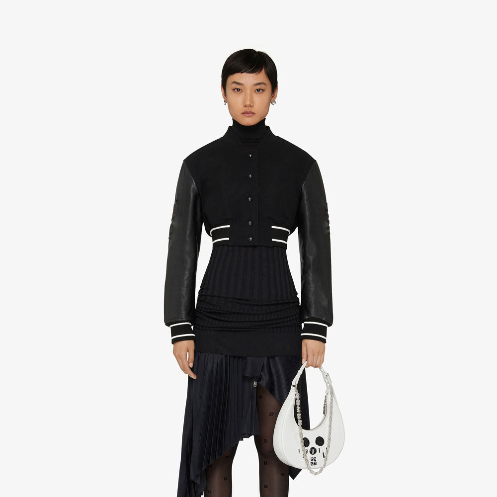 Person wearing a black outfit with leather sleeves holding a white handbag on a white background