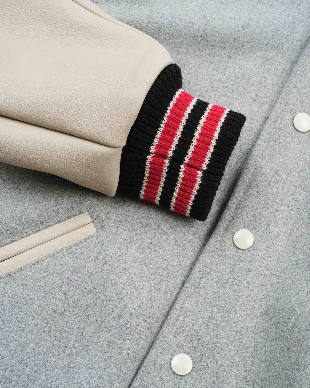 Close-up of a light gray wool-blend jacket with a cream leather sleeve featuring a black ribbed cuff with red and white stripes, secured by white buttons.