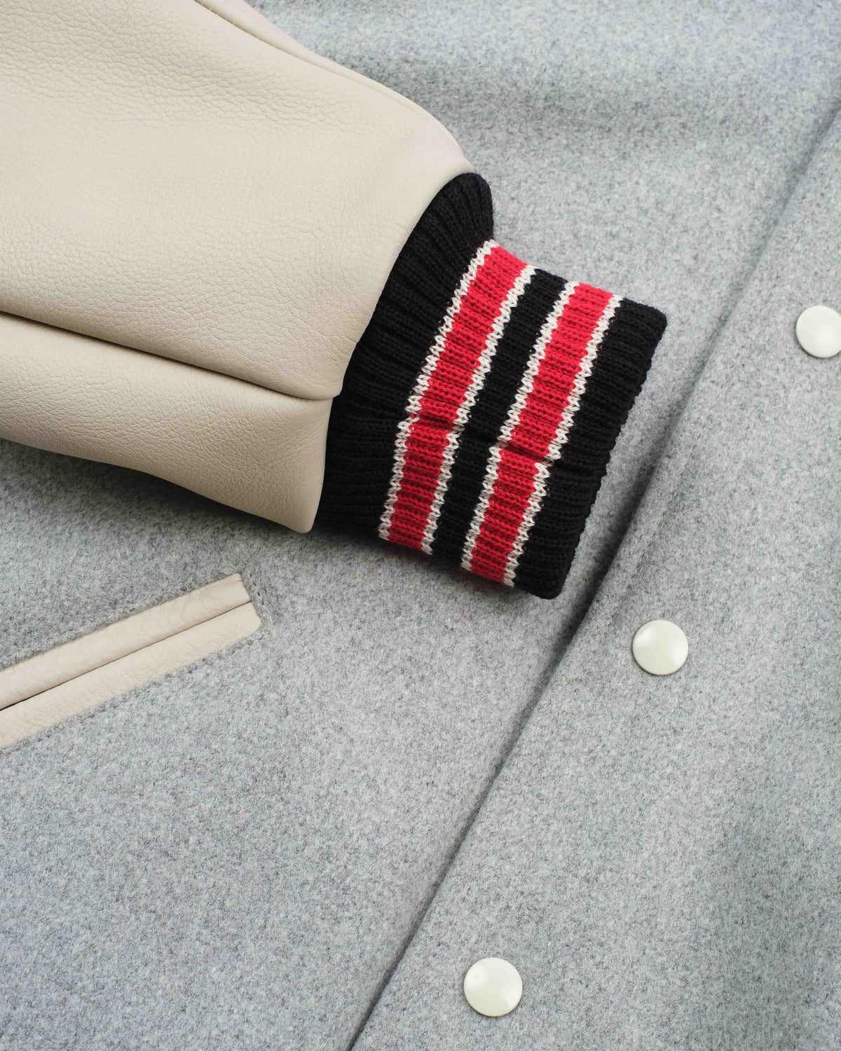 Close-up of a light gray wool-blend jacket with a cream leather sleeve featuring a black ribbed cuff with red and white stripes, secured by white buttons.
