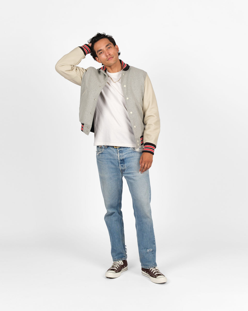A man modeling a varsity jacket with light gray sleeves and cream ribbed collar, cuffs, and hem, worn over a white t-shirt, blue jeans, and brown sneakers against a white studio background.