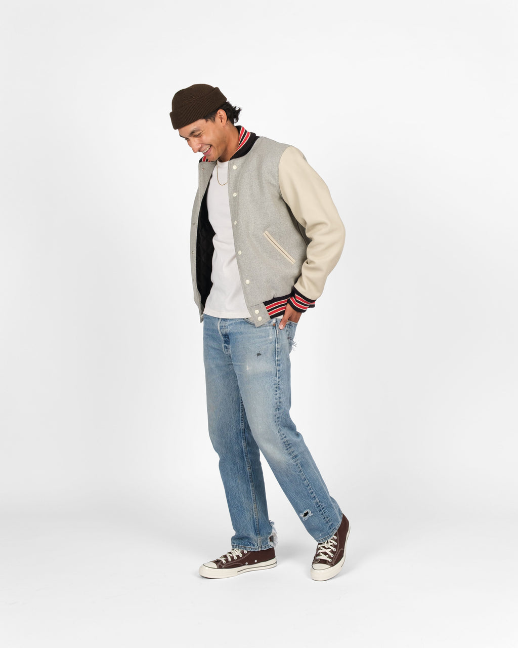 A man wearing a gray and beige varsity jacket, light blue jeans, a white t-shirt, a dark brown beanie, and brown sneakers, standing against a plain white studio background.