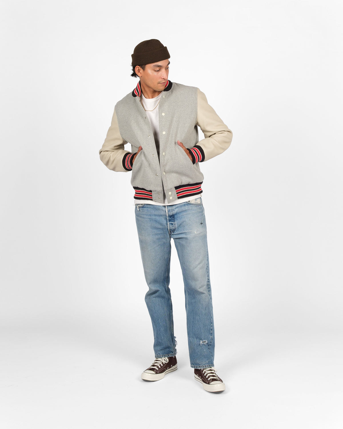 A man modeling a light gray varsity jacket with beige sleeves and red and black striped ribbed trim, paired with light blue jeans, a dark brown beanie, and brown sneakers, against a light gray studio background.