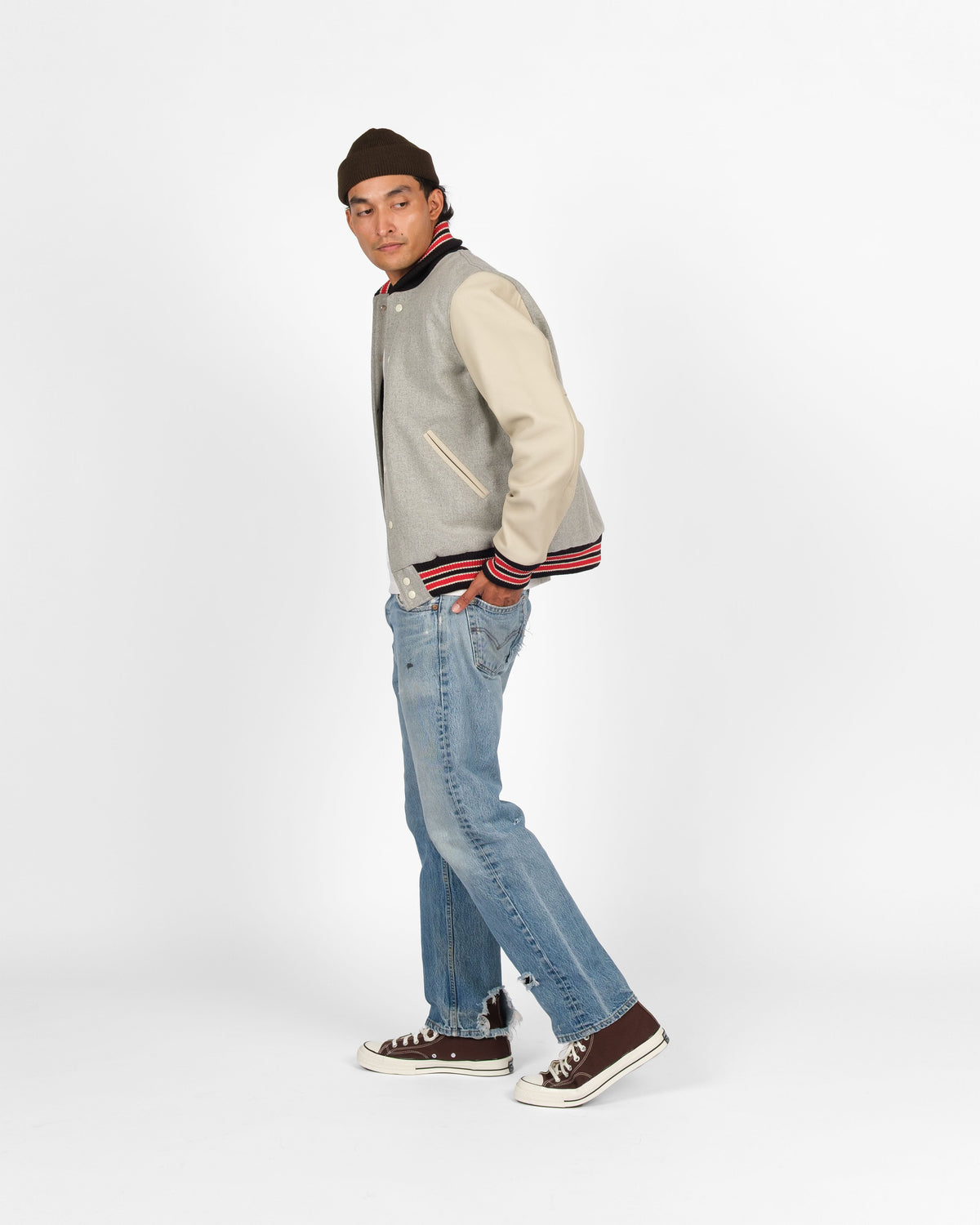 A man wearing a gray and cream varsity jacket, blue jeans, and brown sneakers, standing in profile against a light gray studio background.