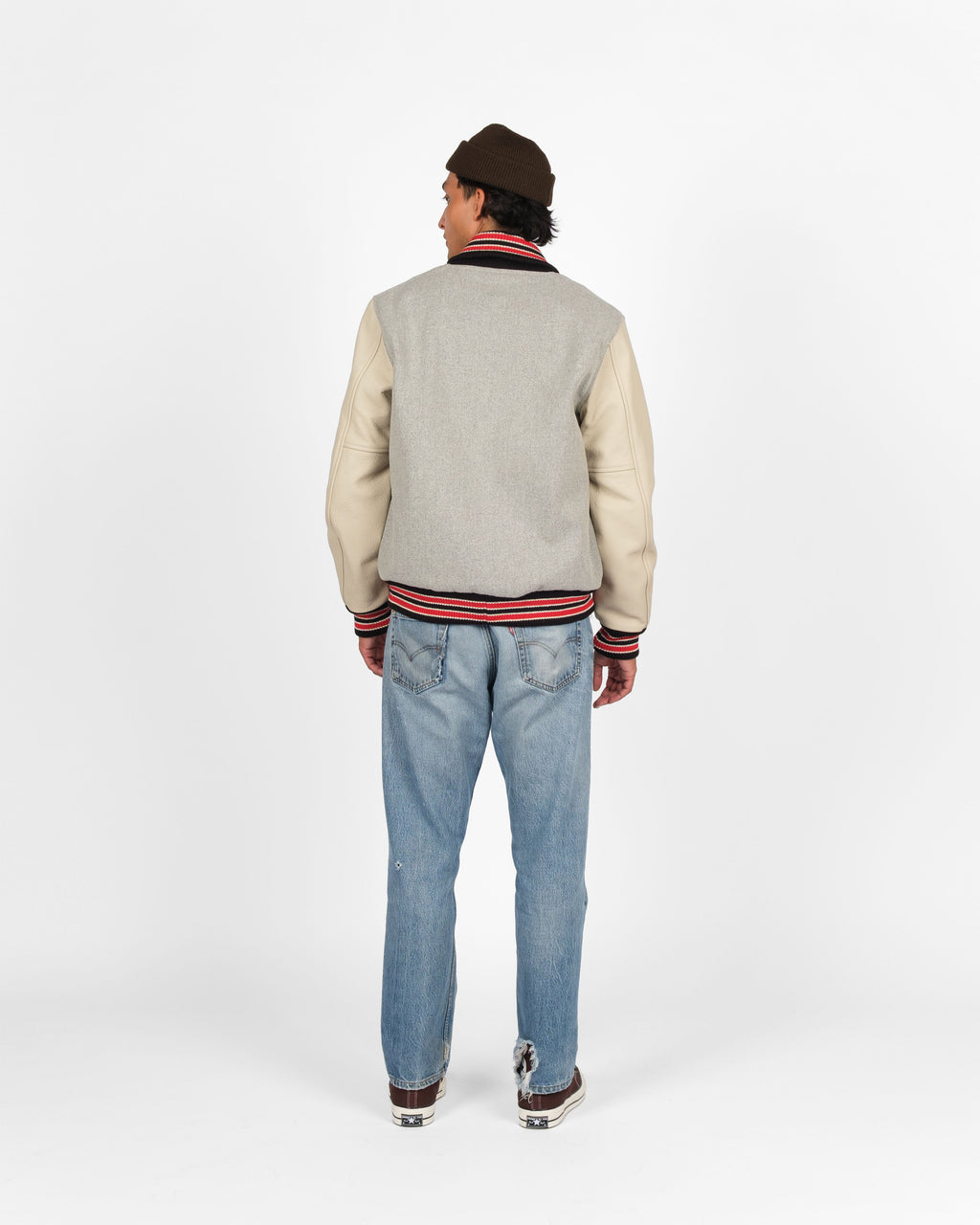 A man viewed from behind wearing a varsity-style jacket with a light gray wool body and cream leather sleeves, paired with light blue jeans, a dark brown beanie, and brown sneakers, against a plain white studio background.