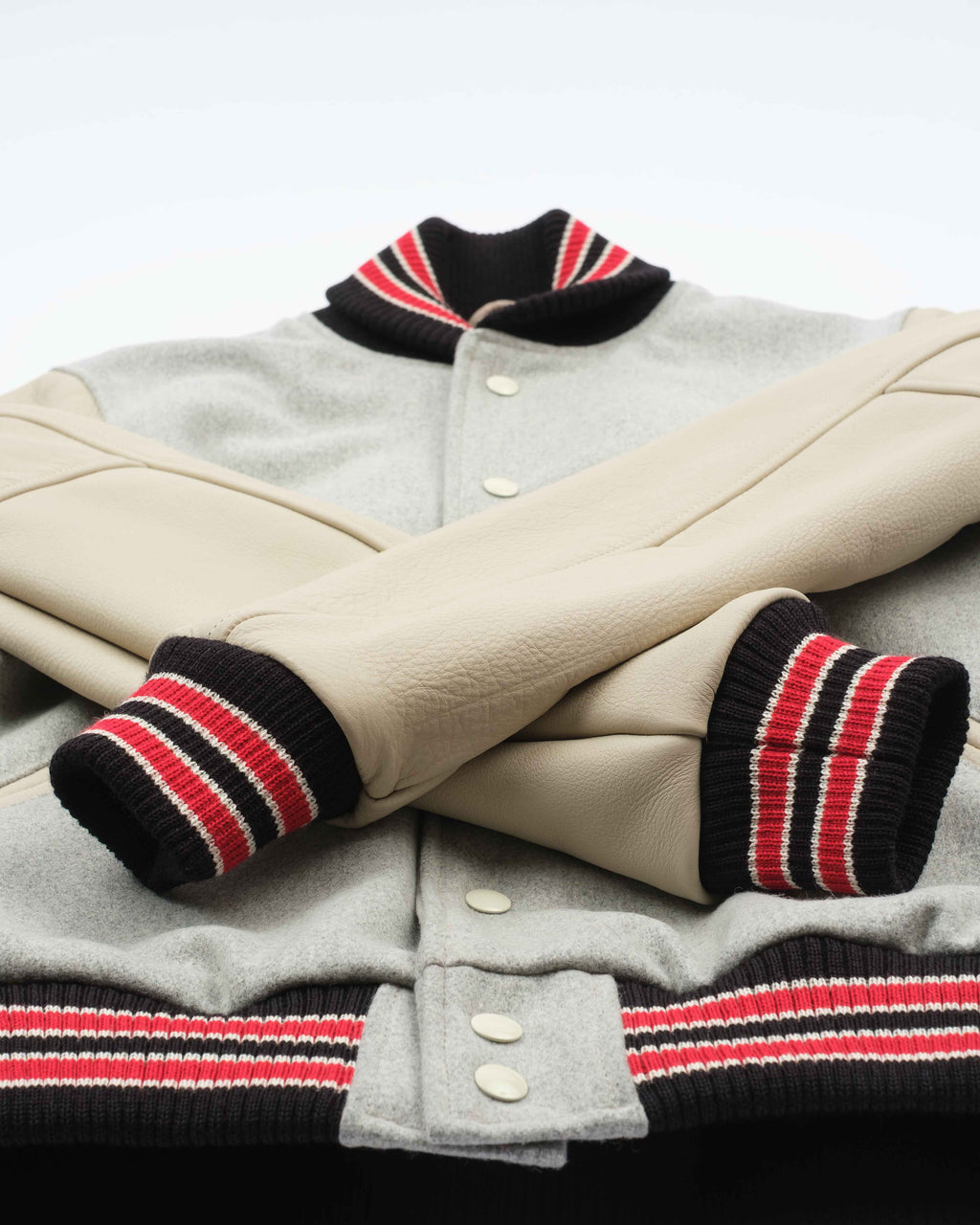 A close-up of a varsity-style jacket with a light gray wool body, beige leather sleeves, and black, red, and white striped ribbed trim on the collar, cuffs, and hem.