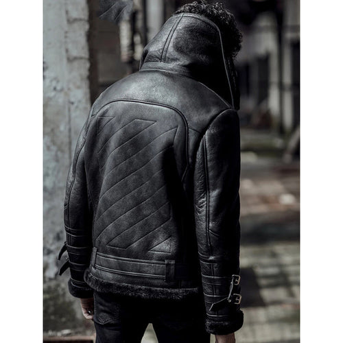 A man viewed from behind wearing a black leather hooded jacket with quilted back paneling and faux-fur trim, standing in an outdoor urban setting.