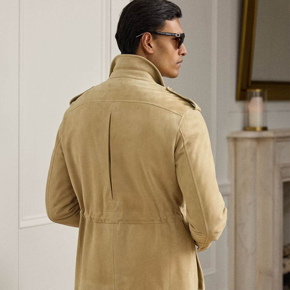 A man wearing a tan suede trench coat and dark sunglasses, photographed from behind in an indoor setting with a cream wall and fireplace visible.