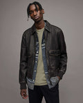 A man wearing a dark brown leather jacket over a patterned shirt and olive green t-shirt, standing against a solid gray studio background.