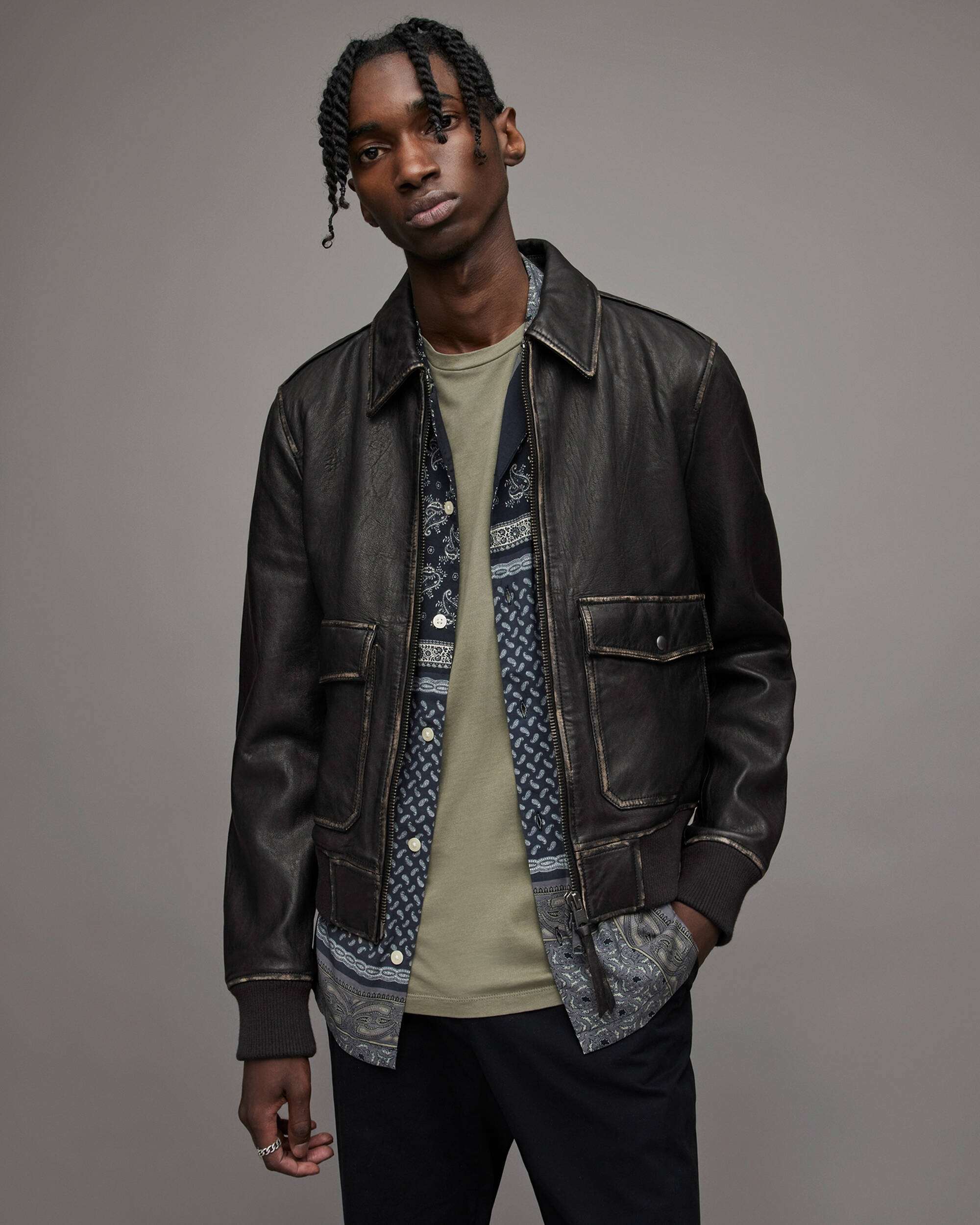 A man wearing a dark brown leather jacket over a patterned shirt and olive green t-shirt, standing against a solid gray studio background.