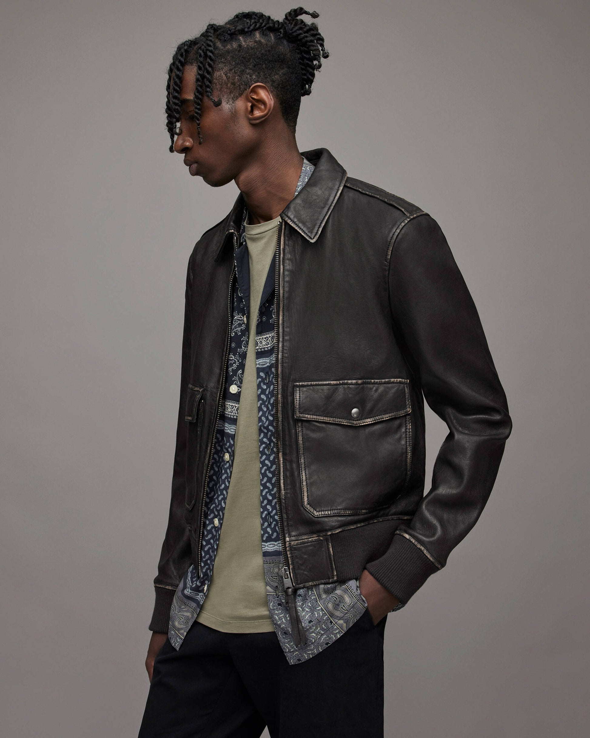 A man wearing a dark brown leather jacket over a patterned shirt and a beige t-shirt, photographed in profile against a solid gray studio background.