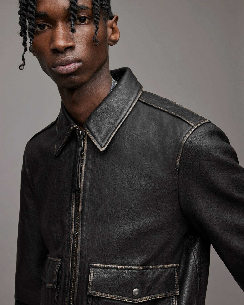 A close-up shot of a model wearing a black leather jacket with contrast stitching, featuring a pointed collar, front zipper, and a chest pocket with a snap button closure, set against a solid gray studio background.