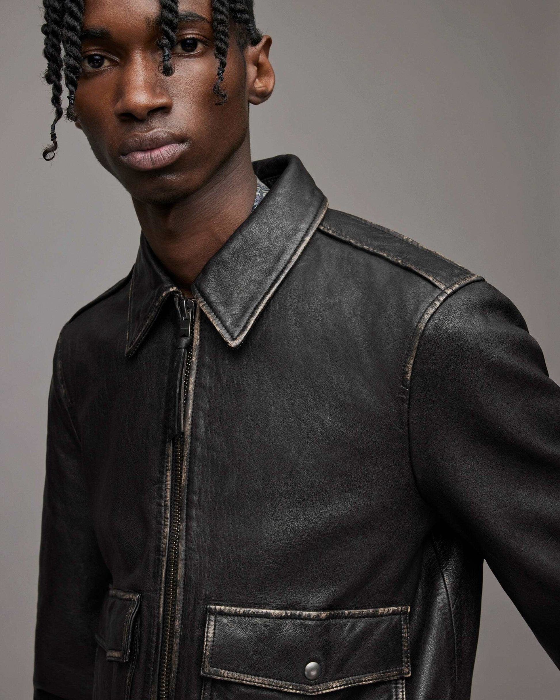 A close-up shot of a model wearing a black leather jacket with contrast stitching, featuring a pointed collar, front zipper, and a chest pocket with a snap button closure, set against a solid gray studio background.
