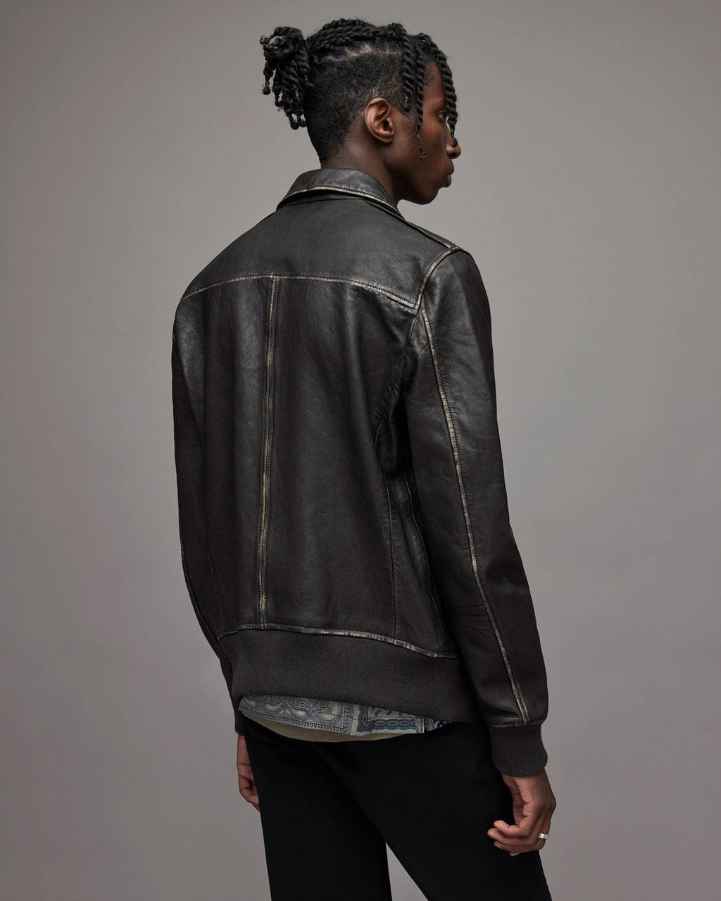 A man shown from the back wearing a black leather jacket with contrast light-colored stitching, paired with black pants, against a solid gray studio background.