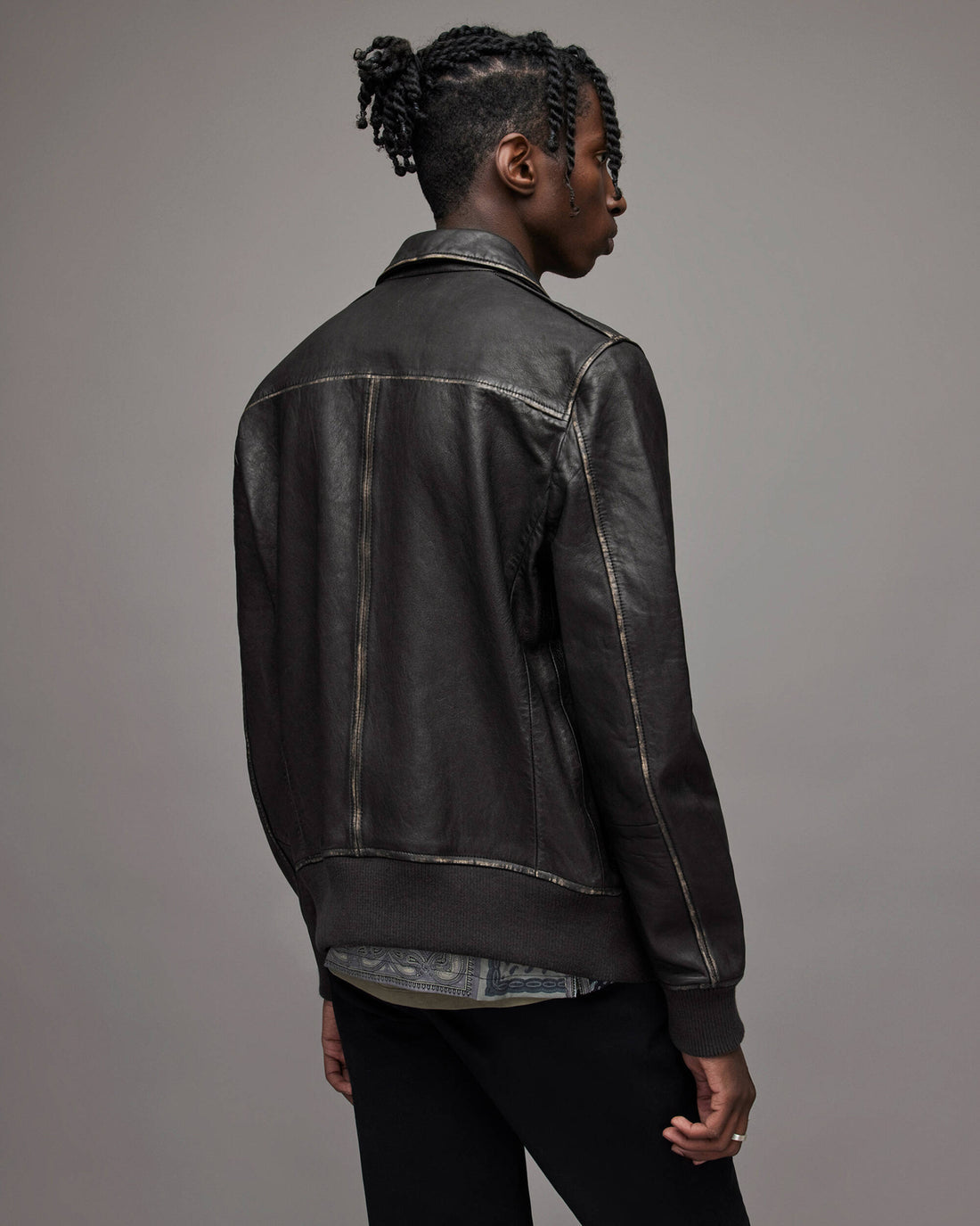 A man shown from the back wearing a black leather jacket with contrast light-colored stitching, paired with black pants, against a solid gray studio background.