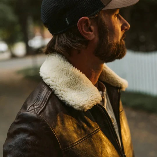 Man wearing a brown leather jacket with a white fur collar and a cap, standing outdoors.