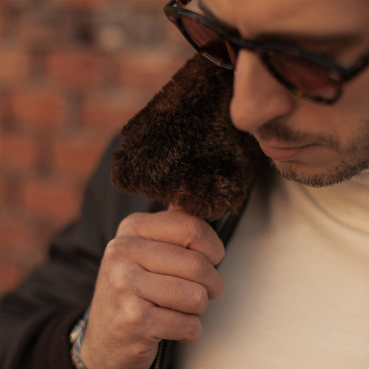 Man wearing sunglasses and a white turtleneck with a brown jacket against a brick wall.